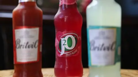 Two out-of-focus bottles of Britvic labelled drink stand next to a bottle of J20 in focus