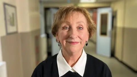 A head and shoulders image of a woman with short dark blonde hair. She is smiling at the camera and is wearing a black jumper with a white shirt underneath and black earrings. 