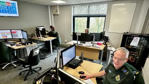 A control room setting with people in green NHS shirts wearing headsets and sitting at desks with computers. On the wall is a screen with numbers and blue boxes.