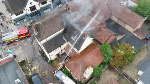 Jayden Stracey An aerial photograph above the Cat and Fiddle pub where a fire has taken place. A hose is being used to put out the fire and smoke is coming from the roof of the building.