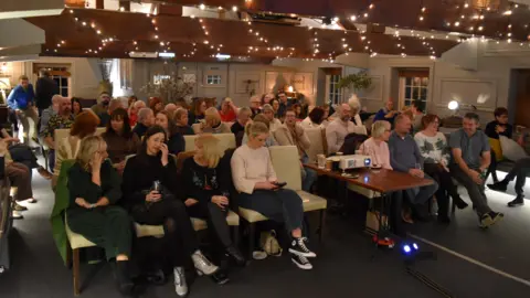Giles Conlon A group of people sat in a warmly lit room. They are all facing the same way. There are fairy lights above the group. 