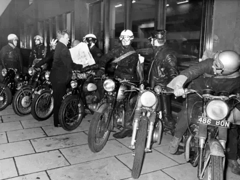 Getty Images A black and white image of half a dozen or so young motorcyclists from the Double Zero Club, Birmingham, lining up on their bikes with newspapers being handed to them