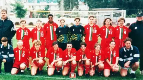 FAW A group photo of the Welsh team in 1995. The players form two rows, with the first row kneeling on the pitch. They are wearing red footballs shirts with a green and white vertical stripe down the middle, and red shorts and socks. Other members of the team and wearing navy jumpers and jackets.