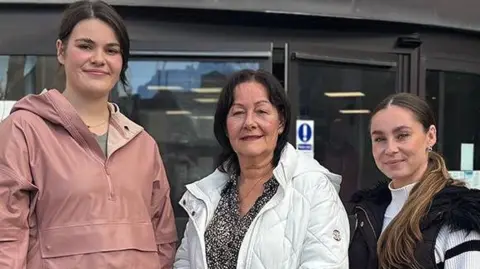 Left to right: Zita with black hair wearing a pink raincoat, Janet with black hair wearing a white coat and black and white patterned top and Natalie with light brown long hair wearing a white and black jumper and black gilet. They are all standing outside the gym and  smiling.