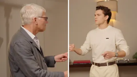 Left, an older man in a suit and glasses points. Right, a younger man in a white shirt points.