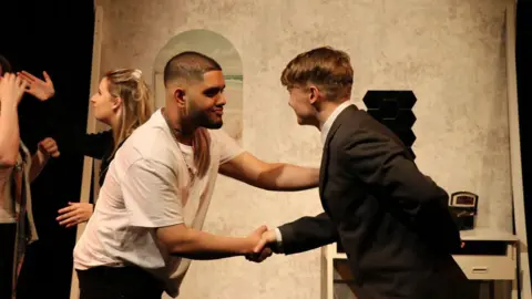 Danyal Zafar A man with short dark hair and a beard in a white t-shirt shaking the hand of a man with blonde hair in a dark suit.