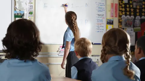 A school girl wearing a light blue shirt and grey skirt, with a long blonde plait, fills in a letter on a white board. Her classmates are watching her complete the word puzzle in their classroom. 