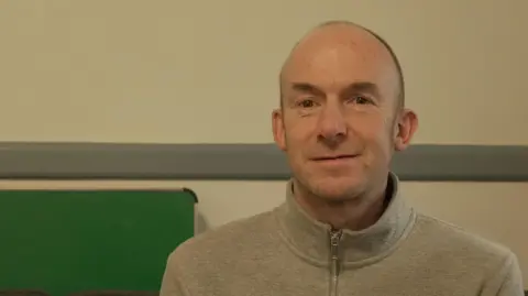 Peter Whittlesea / BBC A man wearing a grey jumper with a zip down the middle. He is stood in a cream room with a green fold-up table leaned against the wall.