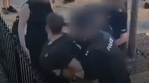 Supplied Slightly blurry CCTV footage showing two police officers behind a young man whom they are trying to restrain up. The young man has a railed metal fence a few inches in front of him. He appears to have his face turned to his right shoulder.