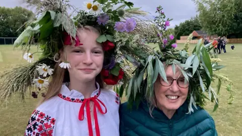 Peter Vacher 17-year-old Alisa, dressed in a traditional Ukrainian outfit, is stood next to her host Polly Vacher on a recreation ground.  Both are wearing garlands of flowers and foilage on their heads.