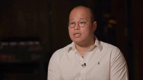 The image shows a man with a bald head and round glasses wearing a white button-up shirt with a small logo on the chest. He is seated and looking slightly off-camera with a neutral to serious expression. The background is dark and out of focus, drawing attention to the man, who appears to be speaking or being interviewed, as indicated by a small microphone clipped to his shirt.