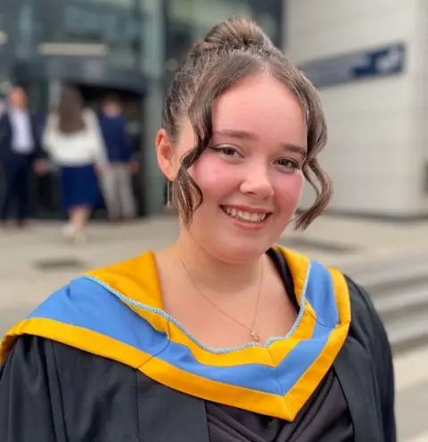 Police Scotland A young woman with her brown hair tied back on graduation day in a black gown with a blue and gold sash around her neck