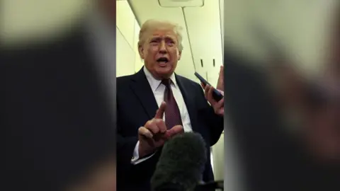 Donald Trump gestures towards a microphone. He is wearing a black suit, white shirt and a dark-coloured tie.