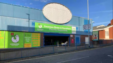 A large warehouse type building alongside a road. A large gree sign over an entrance says Emmaus Home and Furniture Charity Emporium