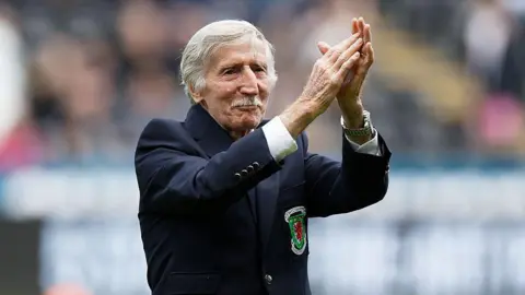 Getty Images Cliff, who has white short hair and a moustache, has his arms raised clapping. He has a blue blazer on with the Wales logo. 