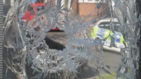 West Midlands Police A close-up image of shattered glass, with a police car and a red vehicle in the background.