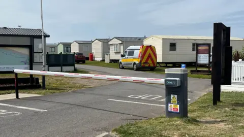 A security barrier bars the road at Redcar Beach Caravan Park. The barrier only covers the road not the grass surrounding it. A row of static caravans can be seen in the park behind it.