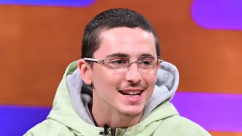 PA Media Timothée Chalamet, who has cropped dark hair and wears glasses, smiles as he appears on sofa of the Graham Norton chat show. He is front of a orange and purple backdrop.