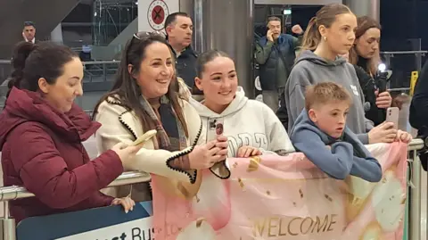 A group of people are standing in an airport waiting to greet family. A young boy and a teenage girl are holding a pink banner that reads 'Welcome home'. 