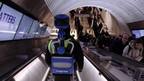 Transport for London A person on the down escalator wearing a Google Maps filming apparatus of a blue rucksack style back pack with a camera poking out on a pole above the bearer's head. Commuters on the opposite escalator look askance at it.