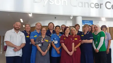 Royal United Hospitals Bath NHS Foundation Trust About a dozen medical staff are posed for the camera in a hospital reception area with a large sign above them saying 'Dyson Cancer Centre'
