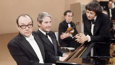 Eric and Ernie sit at a piano while Andre Previn looks on