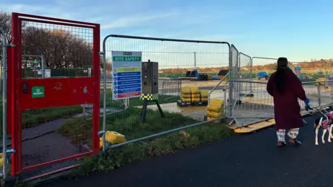Metal barriers with an attached safety sign surround an area containing evidence of construction work. A woman and dog stroll past looking on.