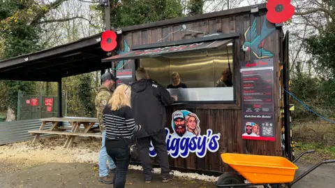 A brown wood shed with Higgsy's written in blue, with people queuing up outside the shed.