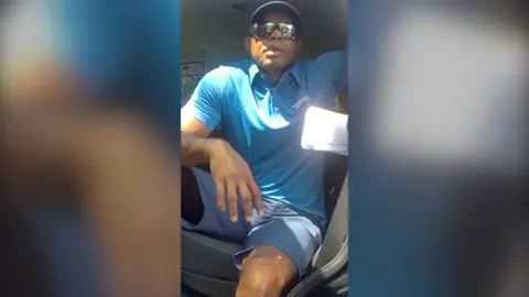 Tiger Woods sits in the driver's seat of his car. He is wearing a cap, sunglasses, slate-grey shorts and light blue polo shirt.