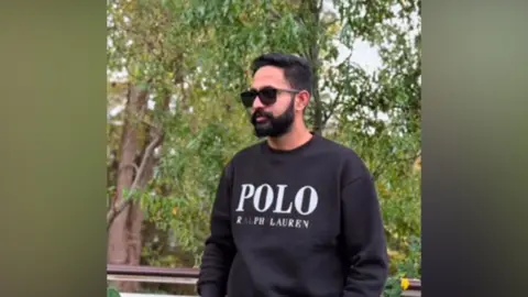 A young man standing outdoors near some trees. He is wearing a dark jumper and sunglasses and has a black beard and black hair. 