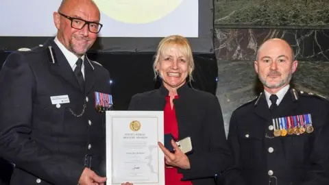 A blonde woman in a black jacket holds a certificate marked "Police Public Bravery Awards". She is flanked by two male officers in uniform