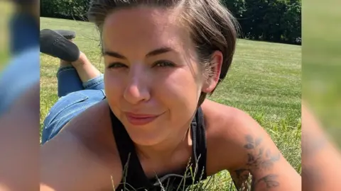 Danielle Moore Photograph of Danielle Moore. She is lying on her front on some grass, with her legs up in the air behind her. She has short, dark hair and is wearing a black vest and jeans. She has a tattoo of flowers on her arm. Her face is turned slightly away from the camera and she is squinting in the sun, but smiling.