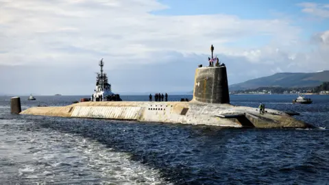 Getty Images HMS Vanguard, a large grey submarine, in the Clyde with sailors standing on top. A tug boat can be seen alongside. 