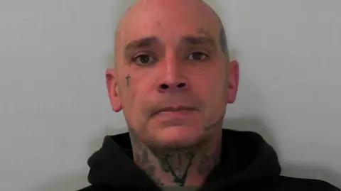 North Yorkshire Police A bald man with tattoos on his face and neck looks at the camera. He is wearing a grey/green t-shirt under a dark zipped-up hoodie.