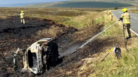 A rural roadside scene where emergency responders are dealing with the aftermath of a vehicle fire. In the foreground, a severely burned vehicle rests on its side in a drainage ditch running alongside a two-lane road. The vehicle's body is heavily damaged and blackened, with much of the exterior burned away, leaving a skeletal metal frame exposed. The ditch contains a small amount of water, and the surrounding vegetation is charred. Two firefighters or emergency workers wearing high-visibility protective clothing and bright helmets are visible in the scene. One emergency worker stands near the road on the right side of the image, directing a stream of water from a hose toward the burned area. The second emergency worker is positioned farther away in the burned field, also using a hose to dampen hotspots.