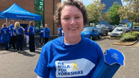 BBC/Seb Cheer A woman, Katie, smiles at the camera. She is wearing a T-shirt which reads "we walk for Yorkshire" and holding a large blue baton with the Yorkshire Cancer Research logo at the top.