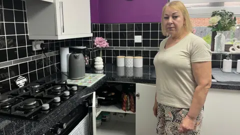 A woman with short blonde hair looking straight-faced and standing in her purple-tiled kitchen in Swindon with white units and black worktops, with the smart meter on view in an open cupboard. 