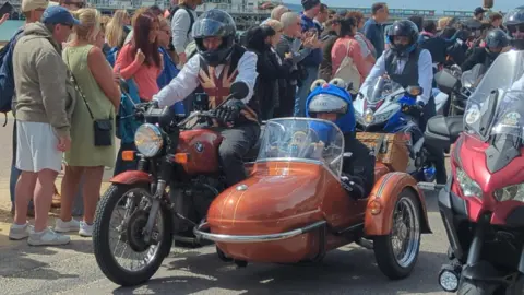 A crows is gathered around an orange-coloured motorbike with a sidecar. The rider of the bike is wearing a black helmet and a Union Jack waistcoat. The passenger in the side car is wearing a hood with cartoonish eyes on the top. A model of Gromit, the dog from Wallace and Gromit, can be seen in the front of the side car. 