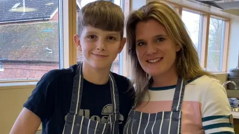 An image of a young boy with brown hair and his mum on his right with blonde hair and a white stripy jumper, both wearing aprons and smiling.
