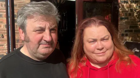 A man in a black t-shirt with grey swept back hair stands shoulder to shoulder with a woman in a red hoodie with shoulder-length red hair. Their faces are in the sun as they stand outside in a garden area. 