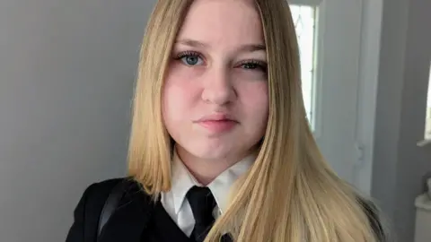 A girl with long blonde hair, wearing a black school jumper, a white shirt and a black tie. 