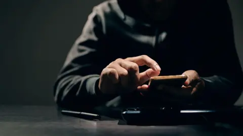 A man wearing a dark hoody has his face concealed. He is holding a mobile phone.