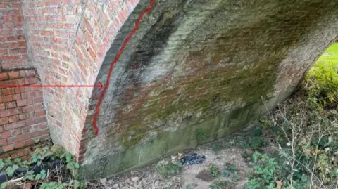 Westwick Group The underneath of a brick bridge has a vertical red line drawn down it. A red arrow has been drawn onto the image towards the red line on the brickwork. 