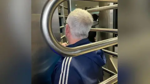 James Bloodworth A man with grey hair faces away from the camera and appears trapped in a metal turnstile.