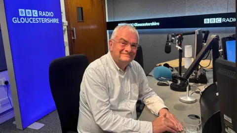 Jeremy Hilton is sitting in a radio studio. He has a half smile on his face, is wearing glasses, has white hair and a white shirt. Two microphones are in shot, one next to the councillor with a blue wind shield and another behind him with a black wind shield. In the background is a door. A lightbox sits to the left of the door which is purple. It has 'BBC Radio Gloucestershire' etched in white type. Another banner which cuts across the wall and is black also has 'BBC Radio Gloucestershire' type etched in white.