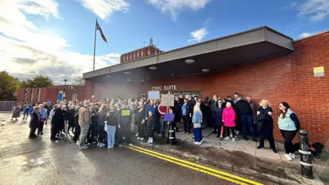 A crowd of protestors stand outside a red-brick building holding up various placards.
