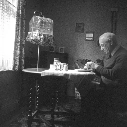 Alec Gill A black and white photograph of an elderly man sitting alone at a small table in a dimly lit room eating a meal. A birdcage with a budgie in it hangs nearby.