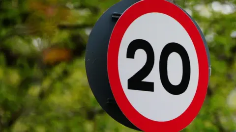 Two round 20mph speed limit signs against a backdrop of green trees - one sign in the foreground and one further away and out of focus.