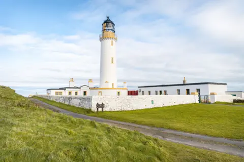 Getty Images A white lighthouse building with yellow windows sits at the end of a pathway through grassland