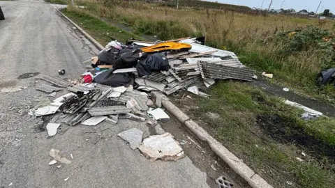 Hull City Council A large pile of fly-tipped waste half on a road and half on a grass verge. The rubbish pile is made up of corrugated roofing sheets and black bags of waste. 
